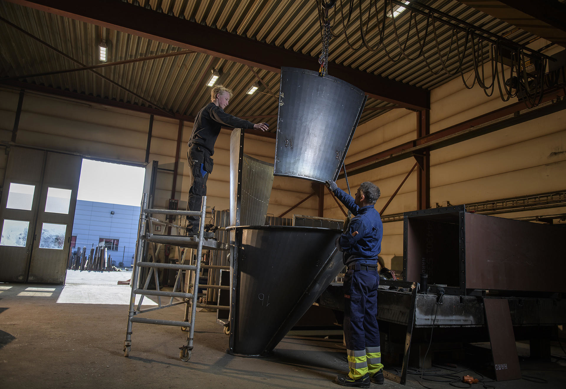 To ansatte i TI Industrier monterer en stor metallkon i ett av våre verksteder.
